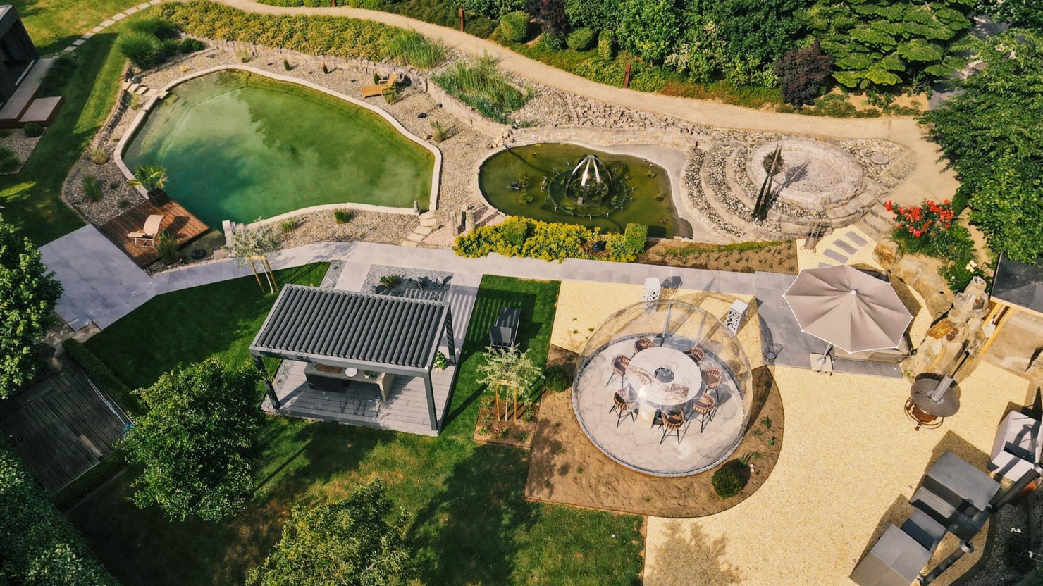 Luftbild des LE Außenshowrooms in Ebensfeld: Naturbadeteich mit Holzdeck und Liege, Wasserbecken mit Fontäne und Steinkreis, Lamellenpergola, gläserne Essplatz-Kuppel, Sonnenschirm, Feuerstelle und Kiesterrasse