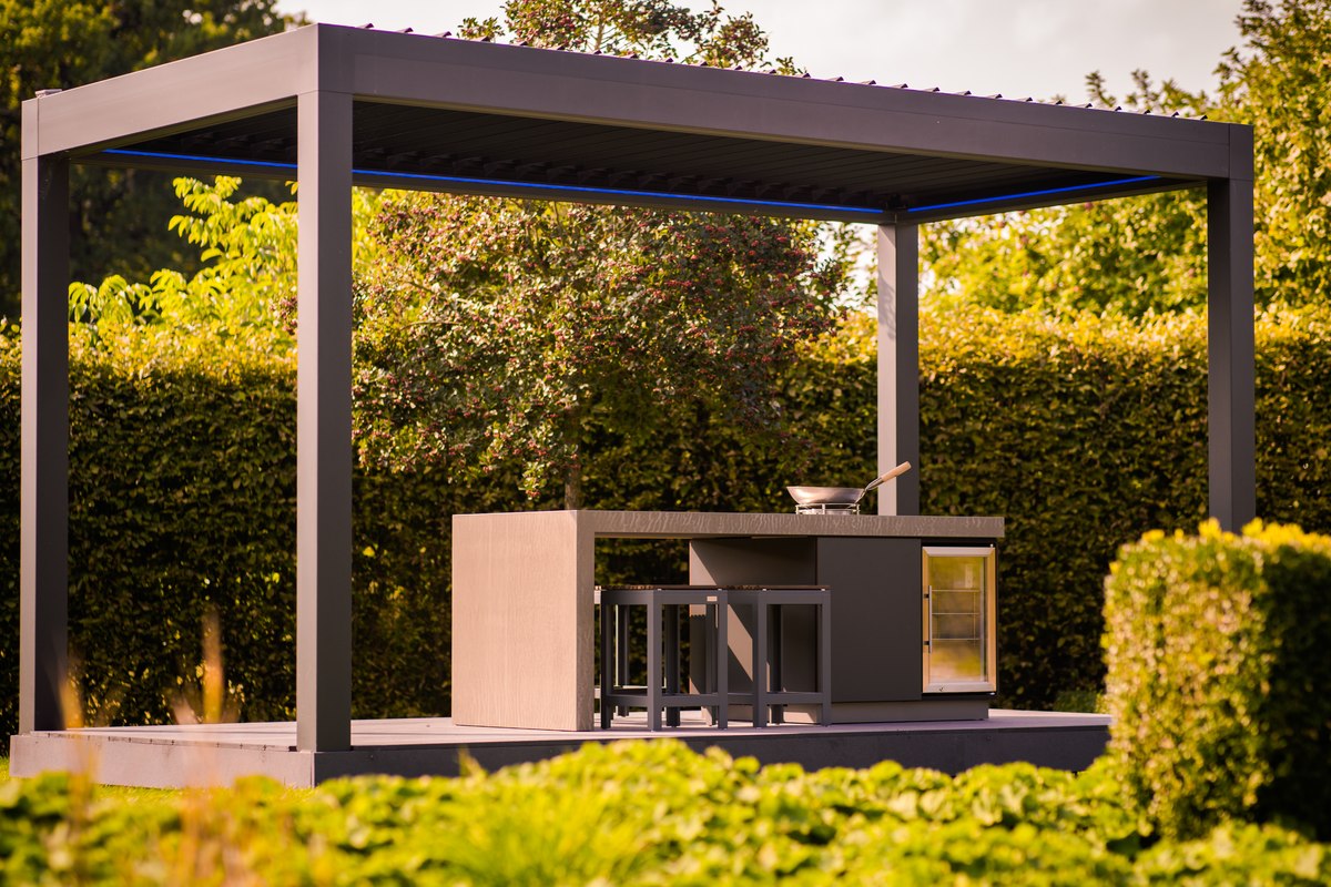 LE KitchenCube unter dem Lamellendach: Outdoor-Küche mit Arbeitsplatte, Wok-Brenner, Glastür-Kühlschrank und Barhockern, darüber das Lamellendach der freistehenden Pergola